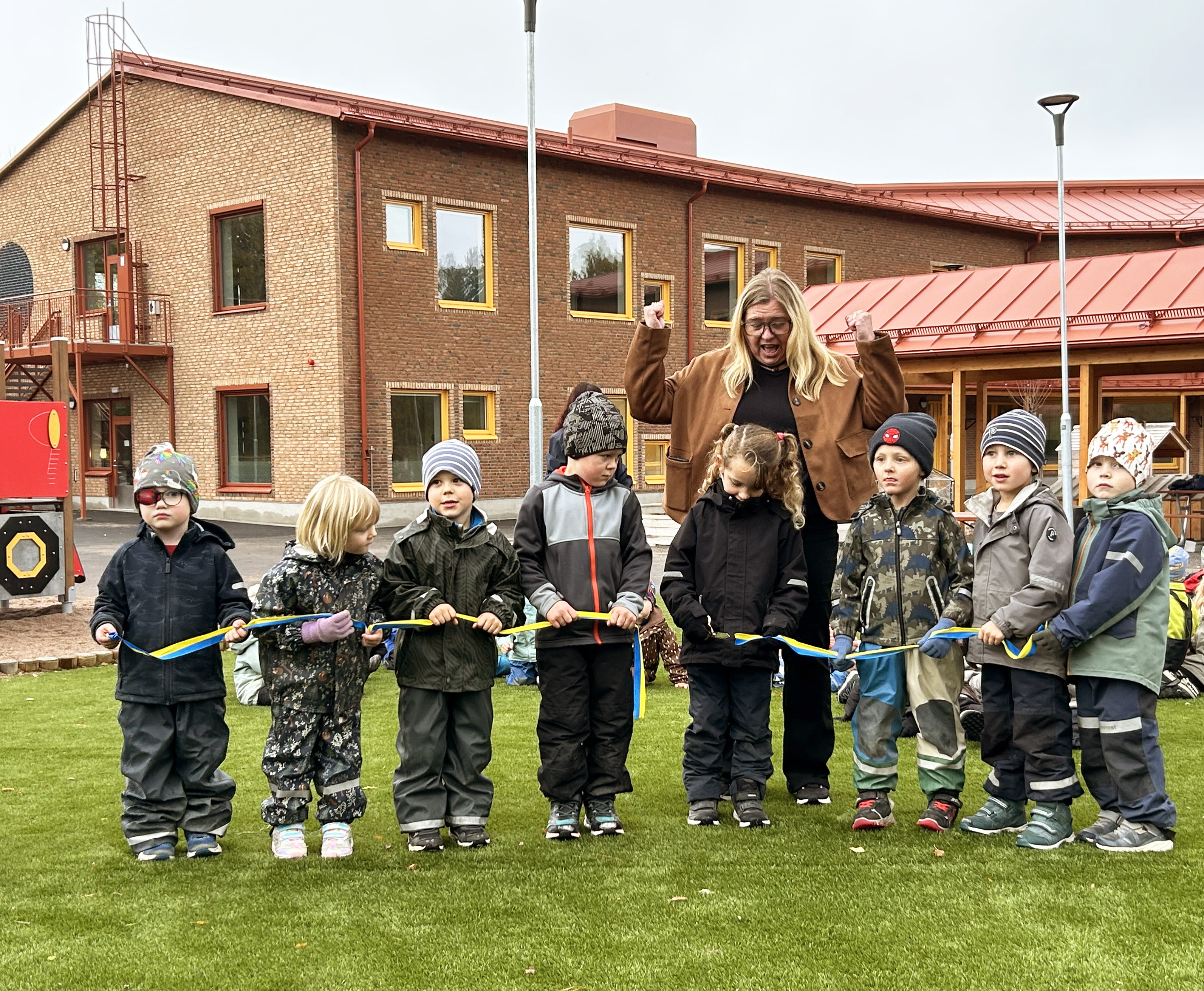 En grupp förskolebarn klipper ett blågult band framför en skolbyggnad, tillsammans med en vuxen.