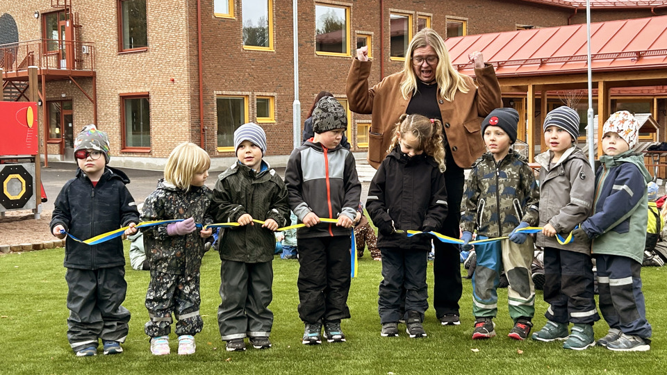 En grupp förskolebarn klipper ett blågult band framför en skolbyggnad, tillsammans med en vuxen.