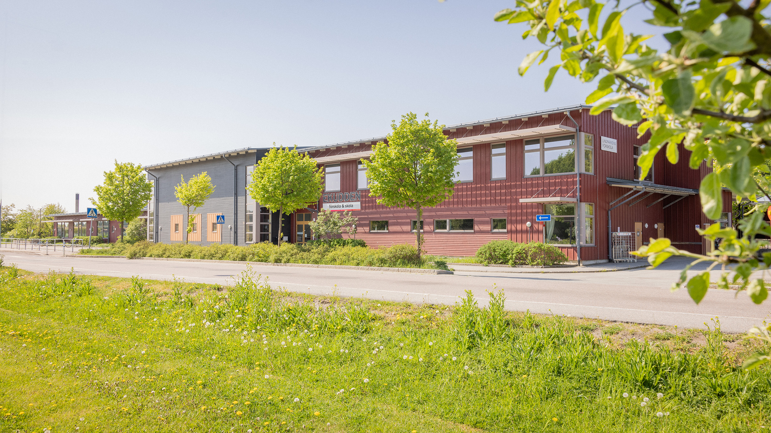 Röd skolbyggnad i trä med träd och bilväg framför.