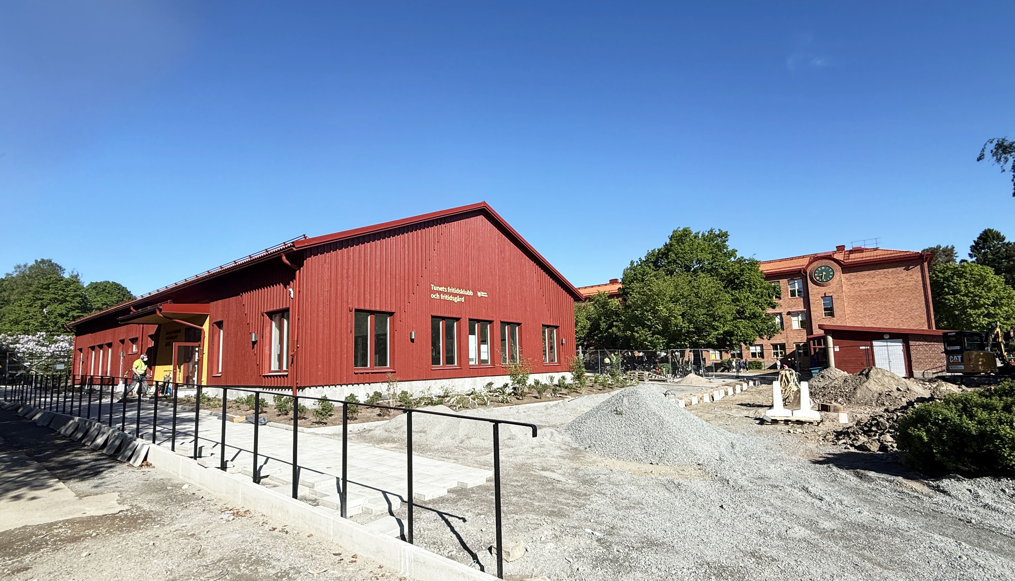 Byggplats där markarbeten pågår framför en ny, röd skolbyggnad.