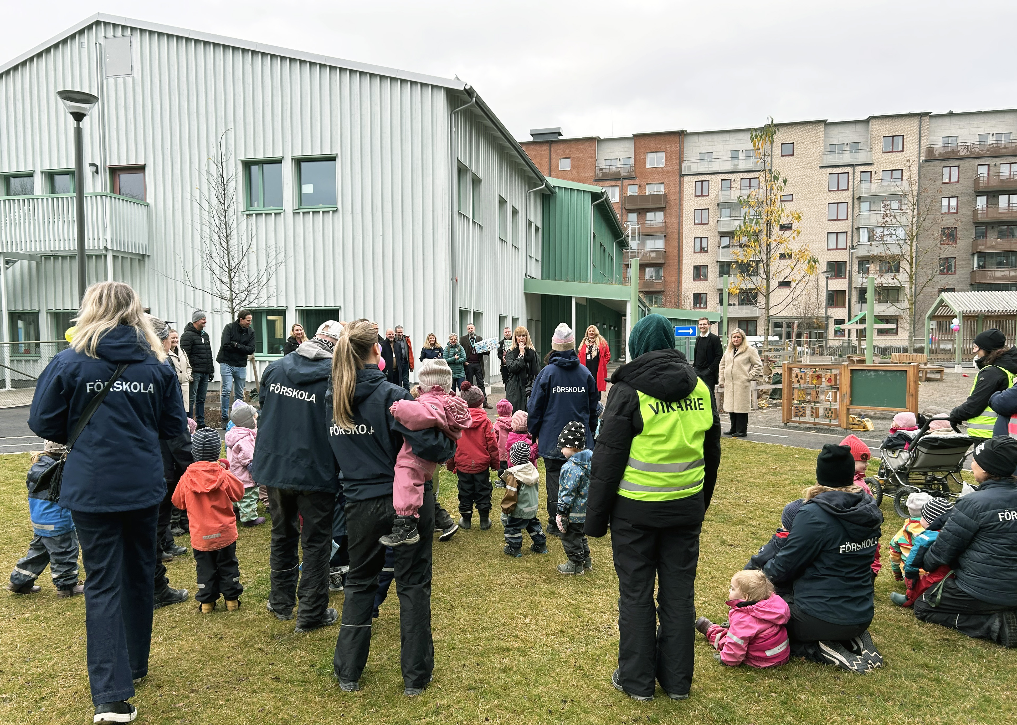 En grupp barn och vuxna står utomhus vid en förskola.