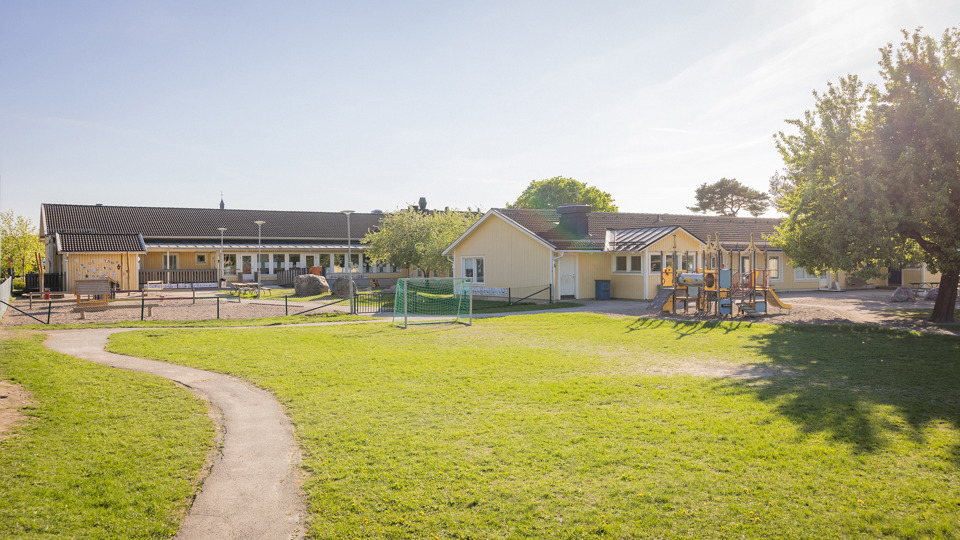 Gul förskola med förskolegård, klätterställning och gröna buskar i förgrunden.