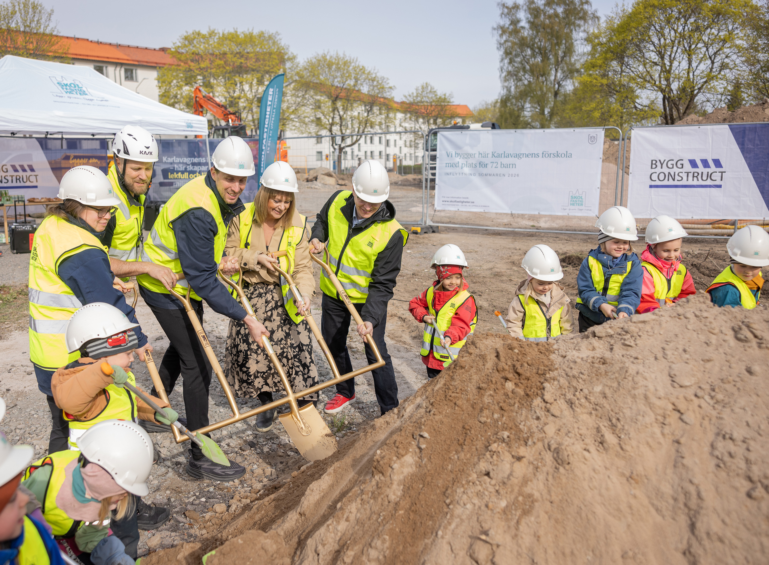Förskolebarn och vuxna i gula västar och vita bygghjälmar gräver i en sandhög på en byggplats.