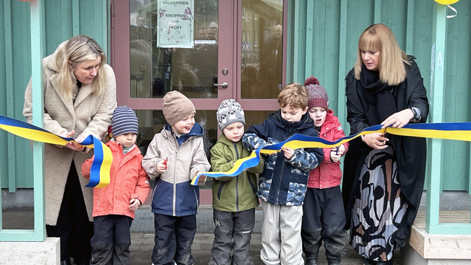 Förskolebarn och två vuxna klipper ett invigningsband framför en byggnad. Det finns ballonger runt dörren.