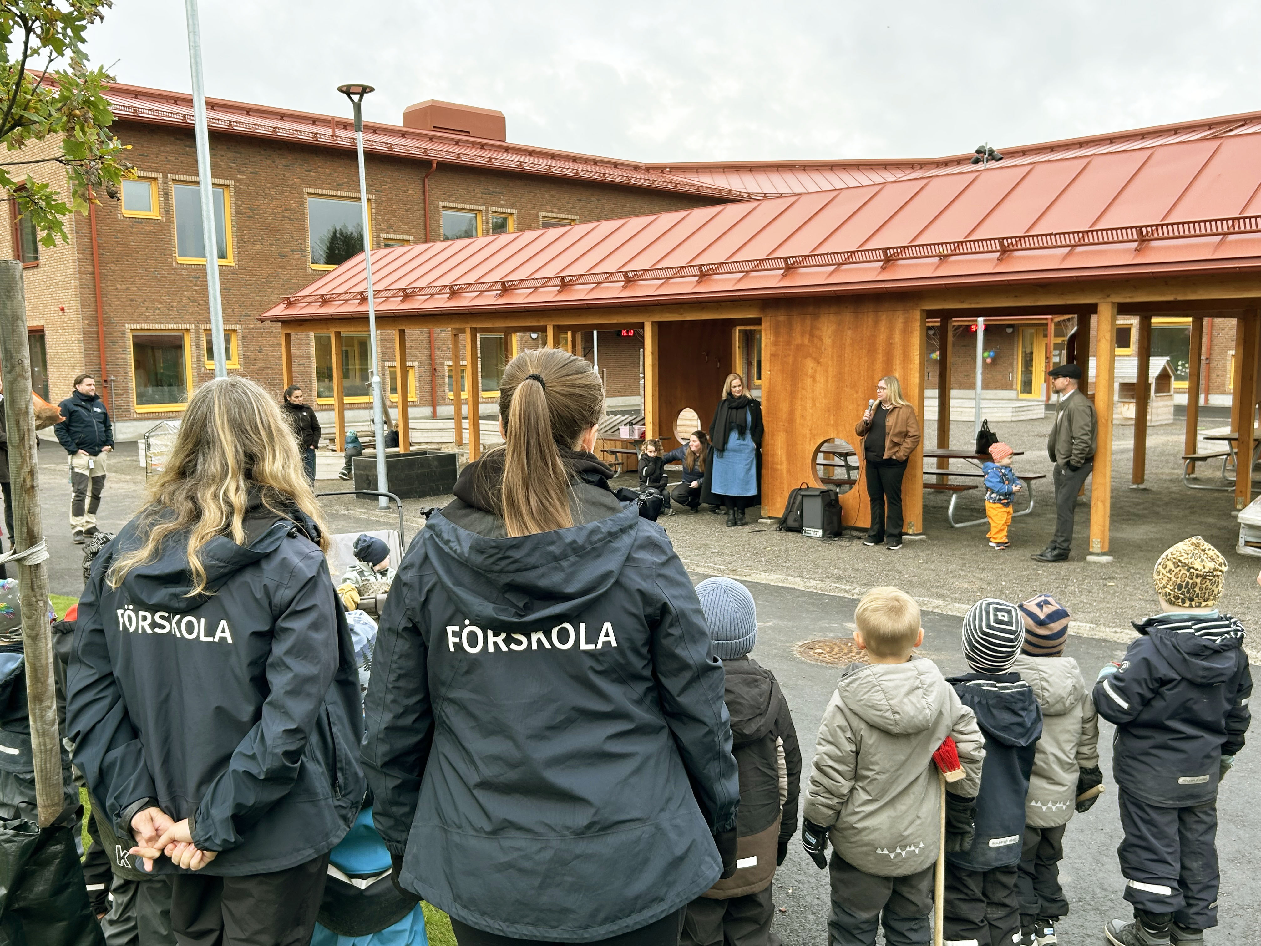 Barn och förskolepersonal samlade på en förskolegård framför en träbyggnad.