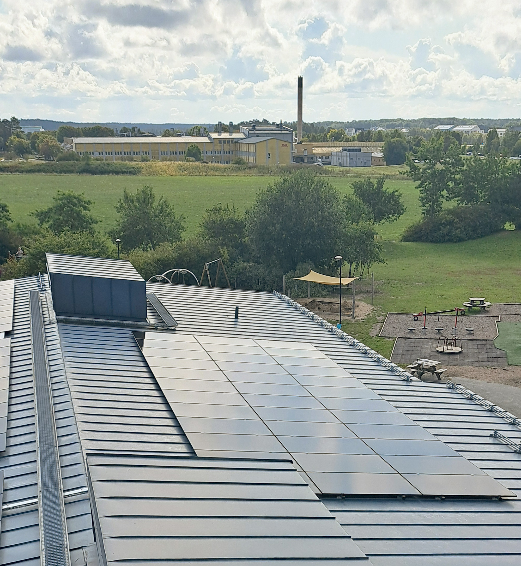 Solcellspaneler på taket av en skola med skolgård och grönska i bakgrunden.