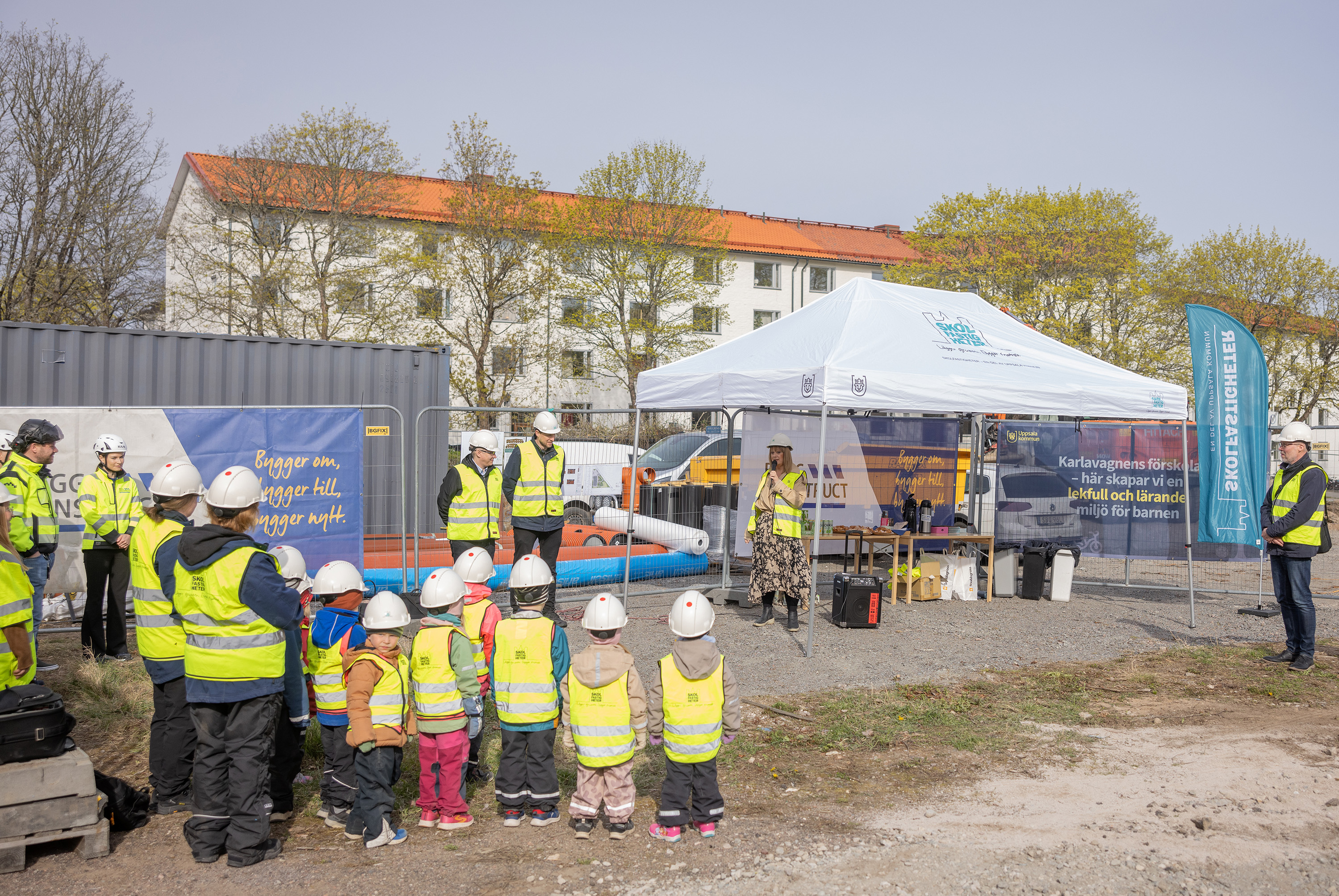 Politiker håller tal framför vuxna och barn i gula västar och vita hjälmar på en byggplats.