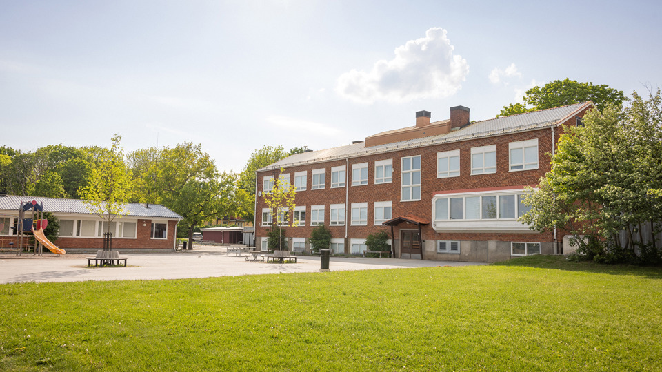 Brun tegelskola med skolgård, träd och rutschkana i förgrunden.