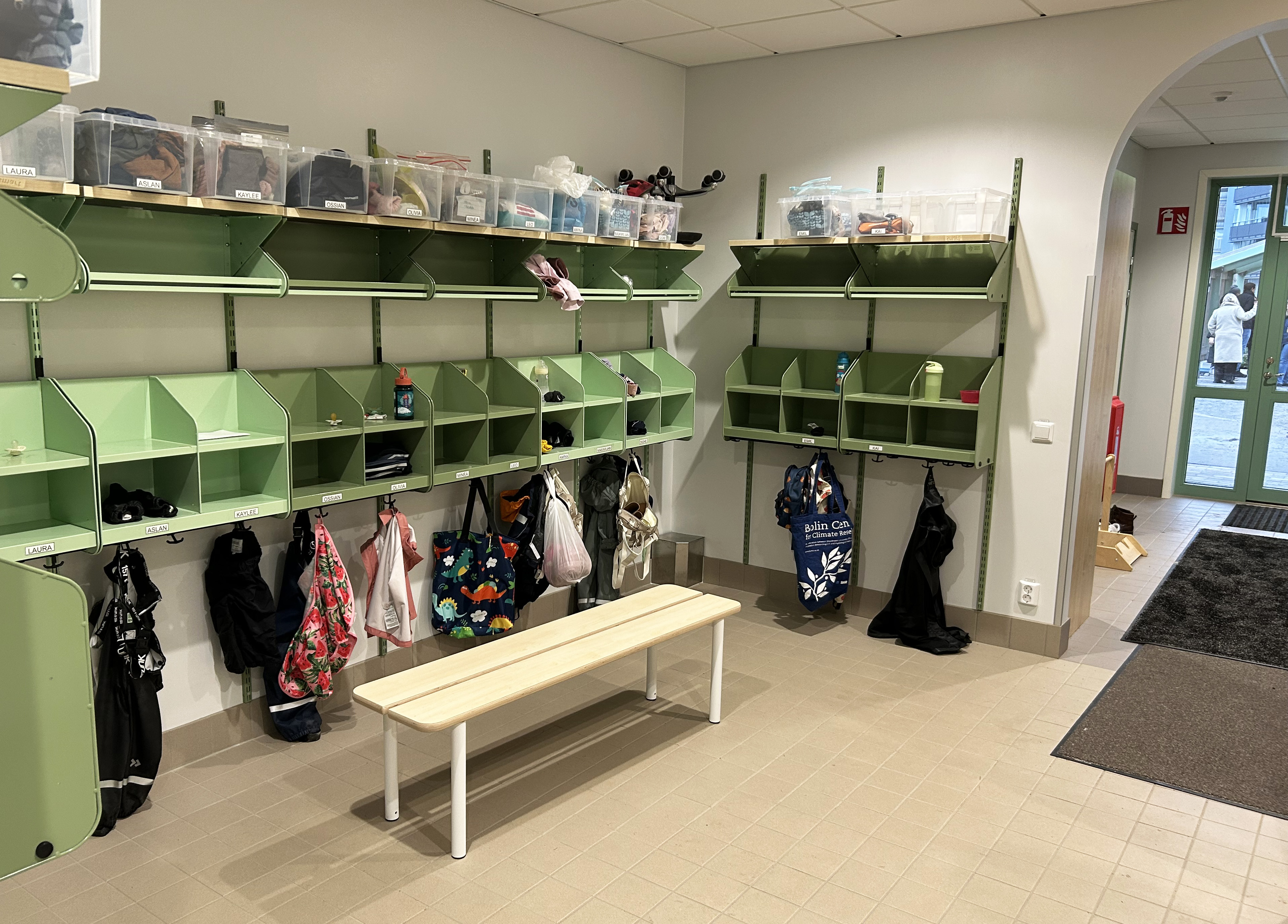 En förskolas entré med gröna fack, klädhängare och plastlådor.