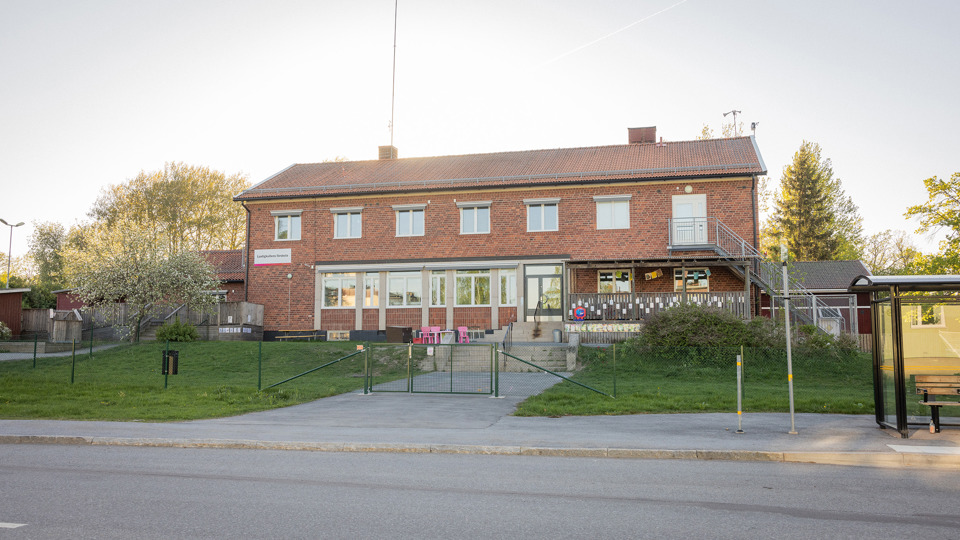 Brun tegelbyggnad med bilväg och busskur i förgrunden.